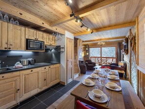 A wooden cabin with a kitchen featuring a microwave, a dining area with a long table set for six, and a cozy seating area with chairs and a fireplace.
