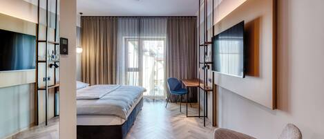 Una habitación de hotel moderna con cama, escritorio, silla, televisor y un amplio ventanal con cortinas.
