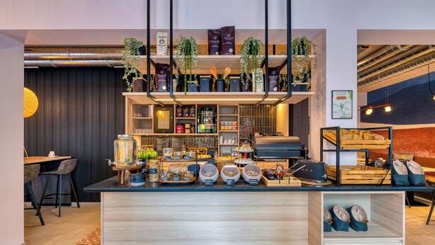 A modern cafe with a wooden counter, shelves with coffee beans and pastries, and a seating area with chairs and a table. - Stadthotel Neumarkt Mitte (Neumarkt in der Oberpfalz)
