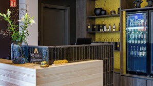 A reception desk with a wooden finish, a glass vase with flowers, a sign reading "Willkommen" (Welcome), and a minibar with various bottles. - Stadthotel Neumarkt Mitte (Neumarkt in der Oberpfalz)