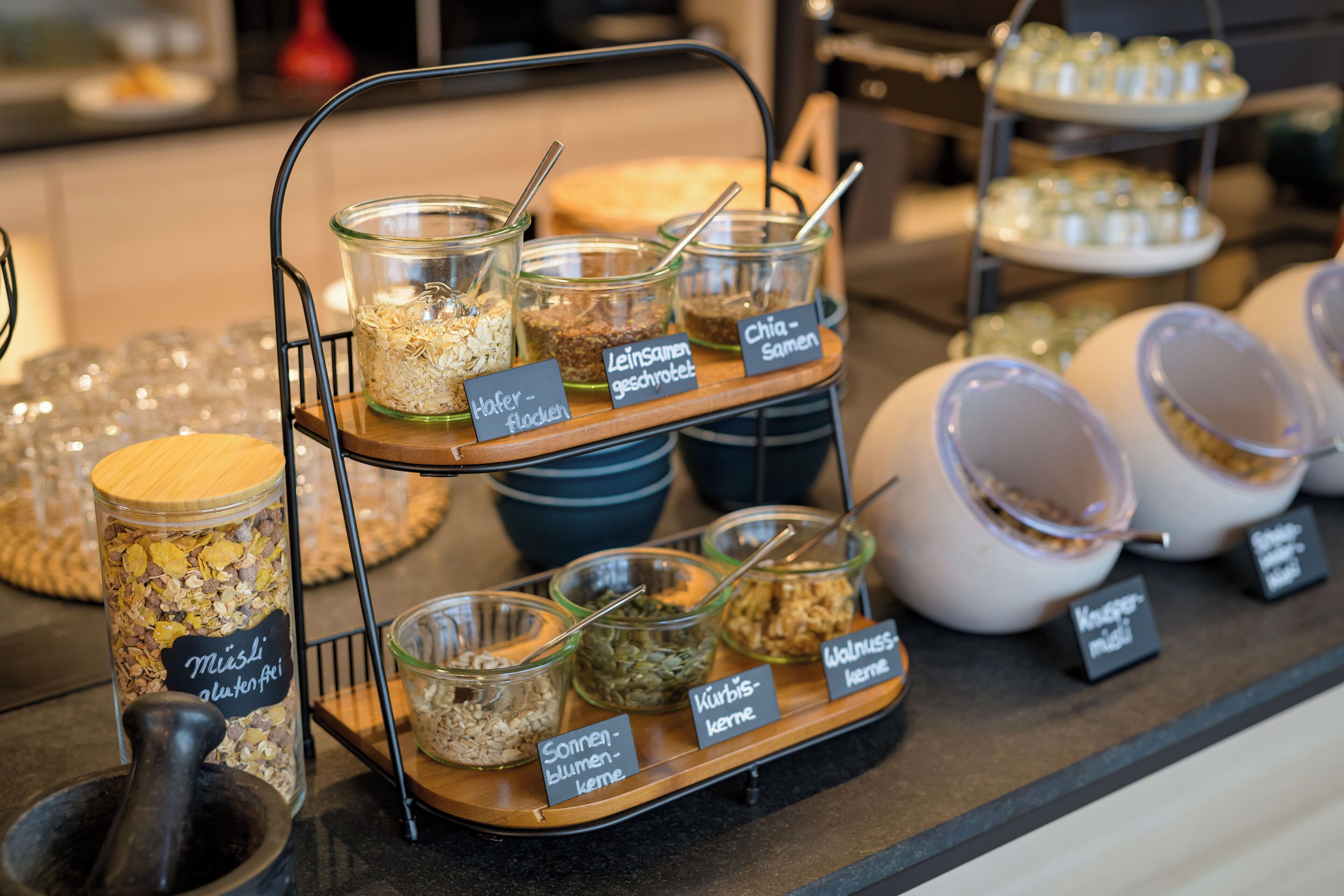 Un bufé con diversos alimentos en recipientes y fuentes de vidrio, etiquetados con nombres en un idioma extranjero.