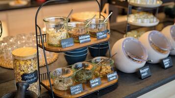 Un bufé con diversos alimentos en recipientes y fuentes de vidrio, etiquetados con nombres en un idioma extranjero.