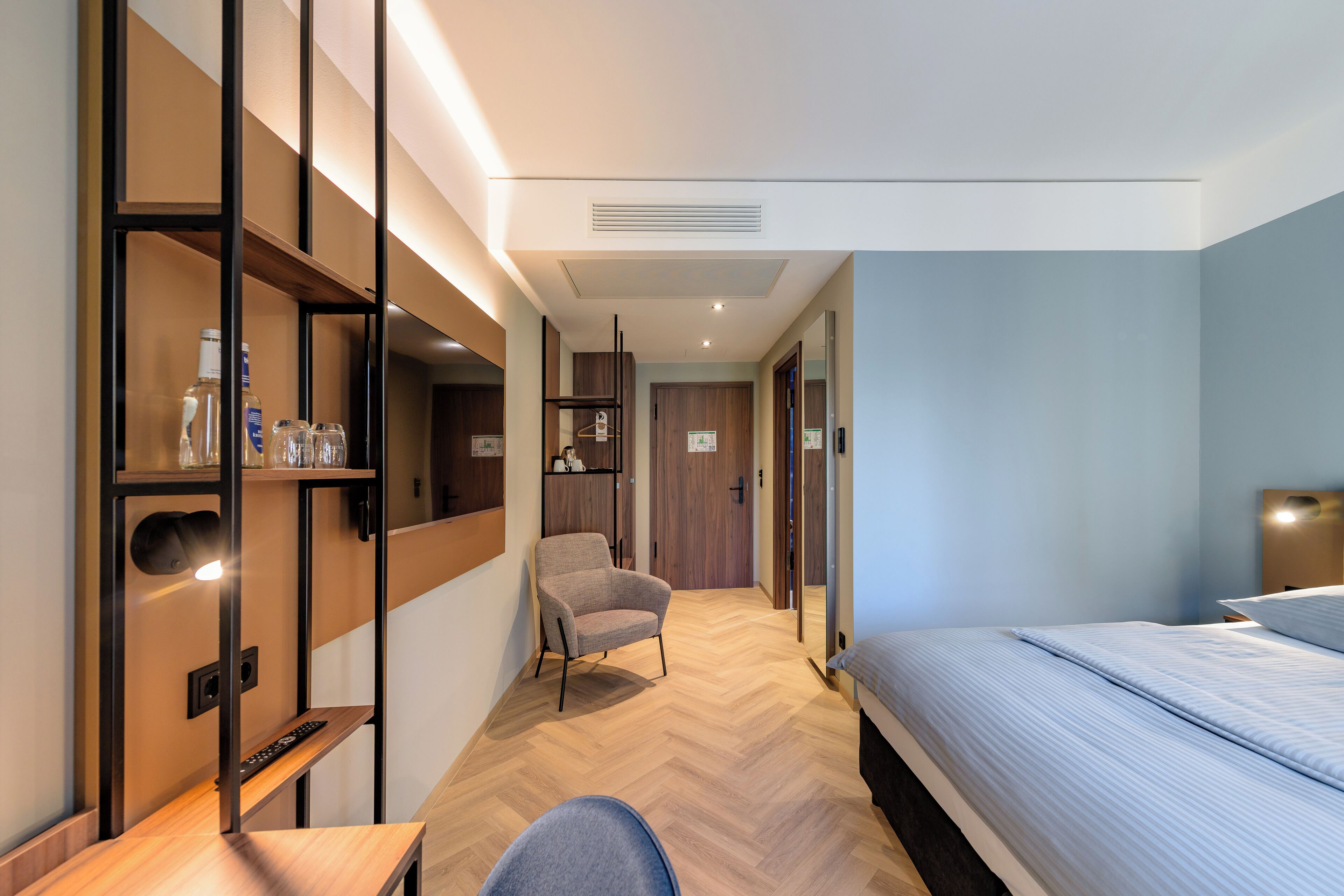 A modern hotel room with a bed, a desk, a chair, and a wardrobe.