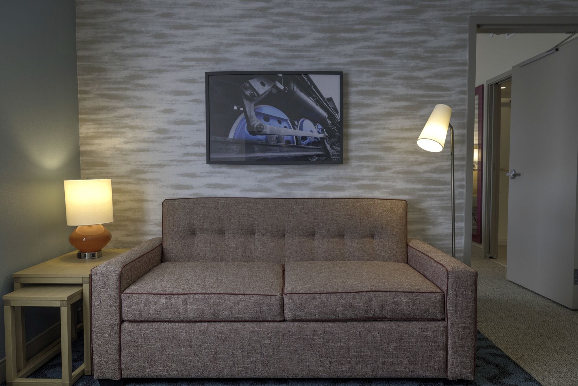 A hotel room with a grey sofa, a framed picture on the wall, and two lamps on side tables.