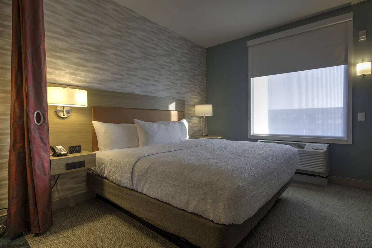Une chambre d’hôtel avec un grand lit, deux tables de chevet avec des lampes, une horloge murale et une fenêtre avec des stores.