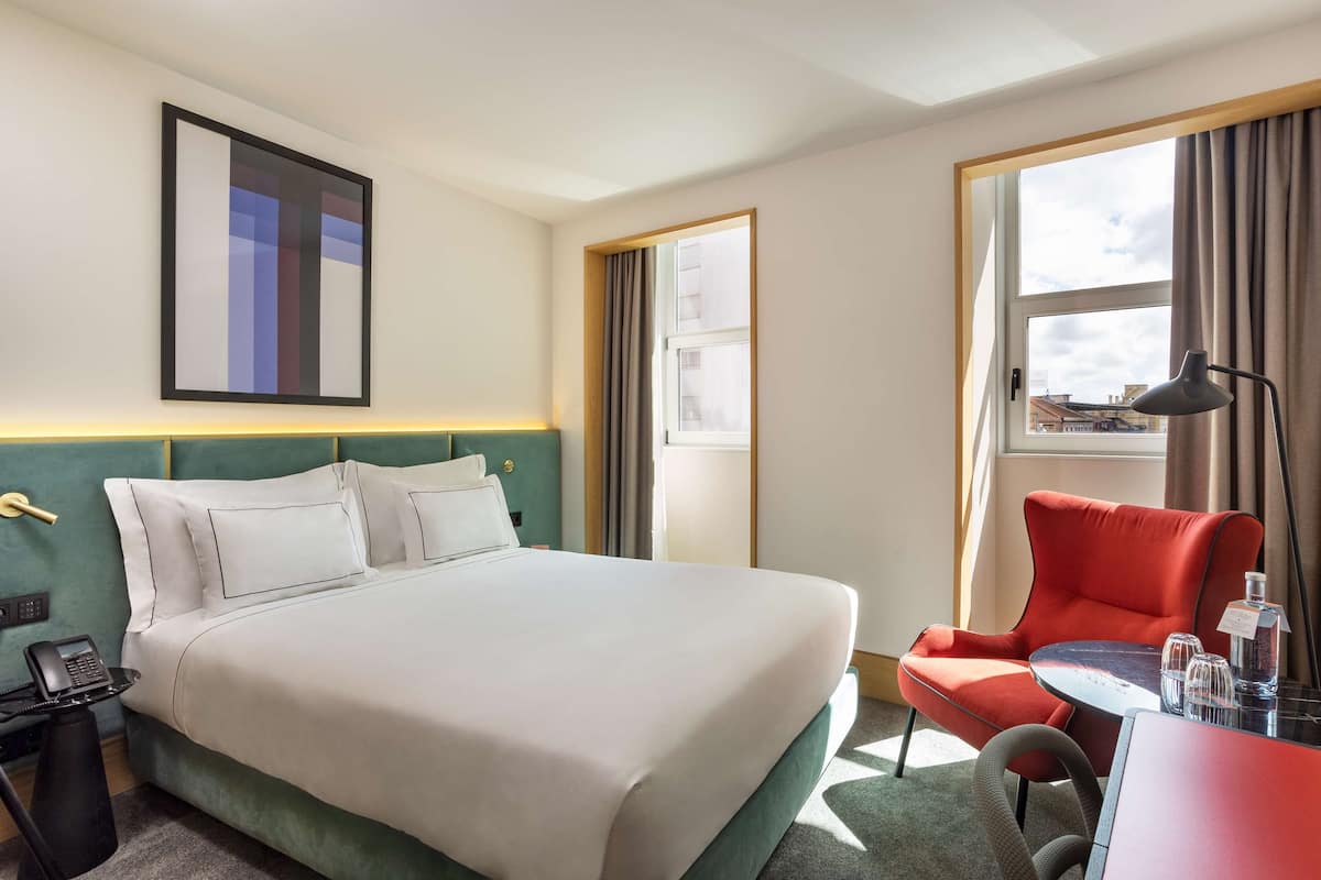 A hotel room with a bed, a desk with a chair, a lamp, and a window with curtains.