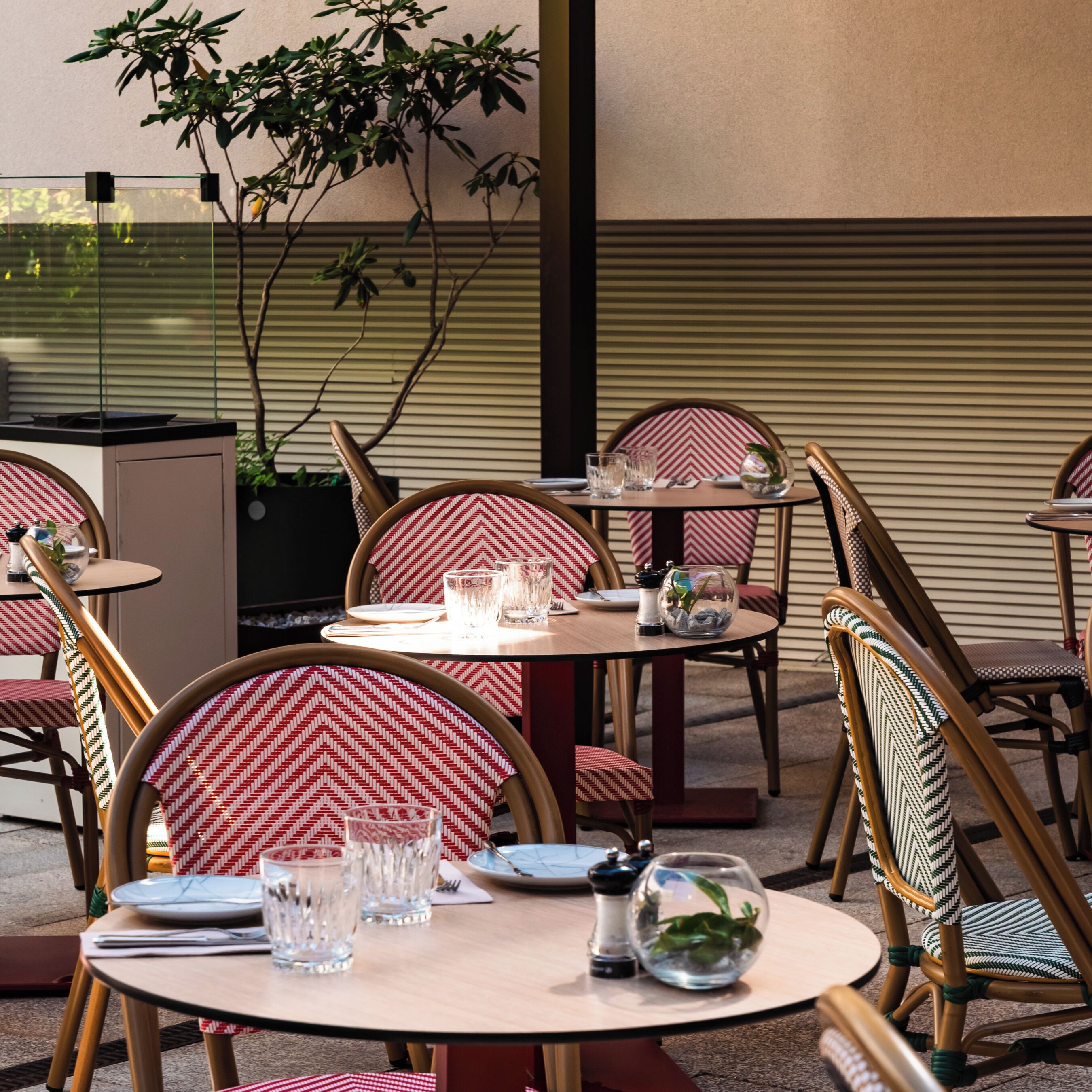 An outdoor dining area with round tables and chairs, some with red and white patterned cushions.