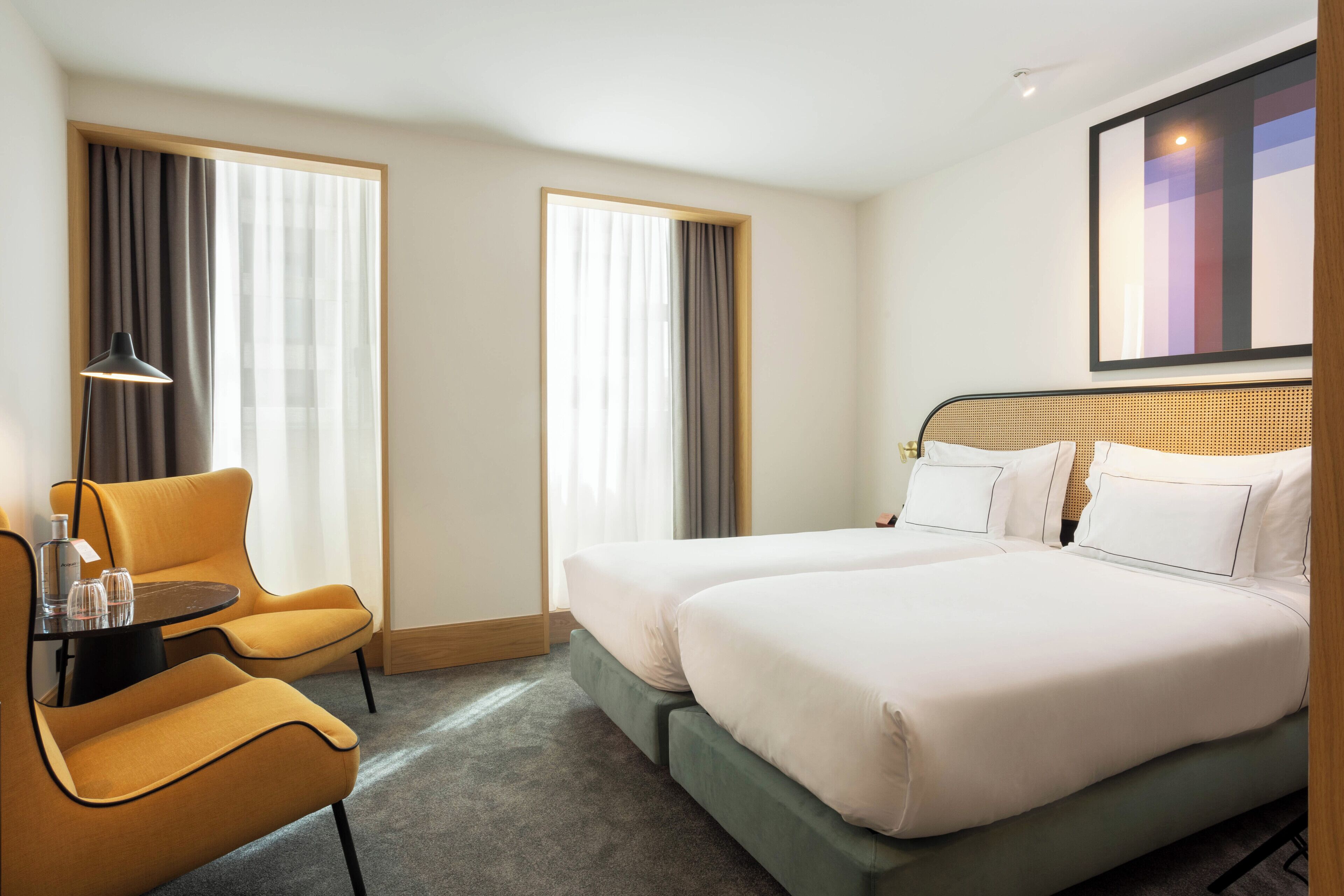 A hotel room with two beds, a desk with a chair, a lamp, and a painting on the wall.
