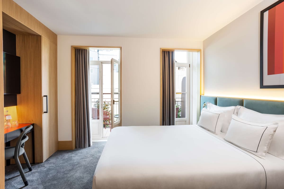 A hotel room with a large bed, a desk with a chair, a TV, and a balcony with curtains.