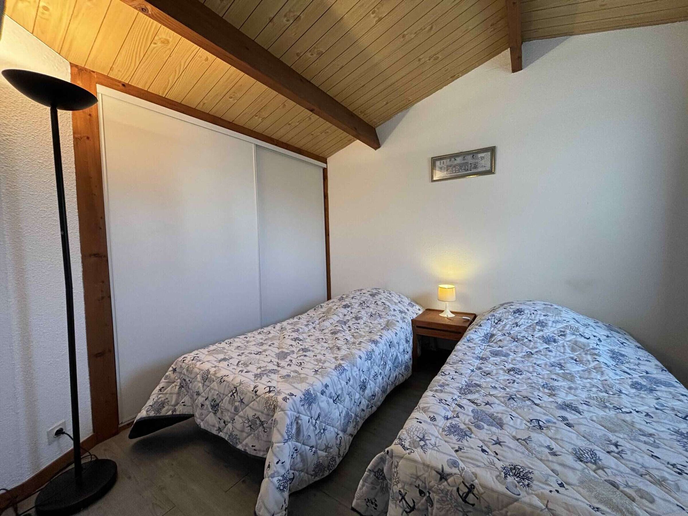 A bedroom with two beds, a bedside table with a lamp, and a wooden ceiling.