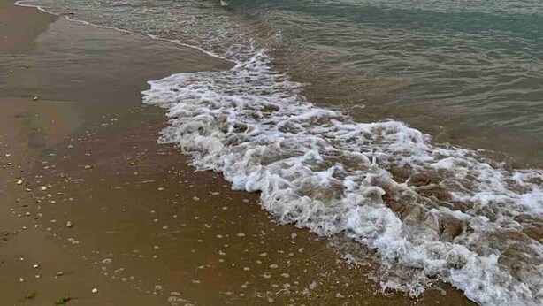 Ein Strand mit sanft anschwellenden Wellen, ein weiter felsiger Ausläufer im Hintergrund und ein klarer Himmel.