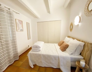 Een netjes opgemaakt bed met witte lakens en een houten hoofdbord, een klein nachtkastje met een witte kaars, en een raam met een gestreept gordijn.