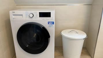 Una lavadora de carga frontal con pantalla digital, un bote de basura blanco y una pared de azulejos.