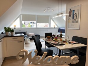 Une cuisine moderne avec un coin repas, agrémentée d’une pancarte « Bienvenue », d’une table dressée avec des assiettes, d’une bouteille de champagne et d’un téléviseur en arrière-plan.