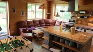 Un chalet en bois accueillant, avec un espace de vie comprenant un canapé en cuir, une table de baby-foot et une cuisine dotée d’un grand îlot central.