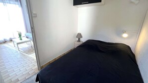 A bedroom with a bed, a bedside table with a lamp, a window with curtains, and a wall-mounted air conditioning unit.