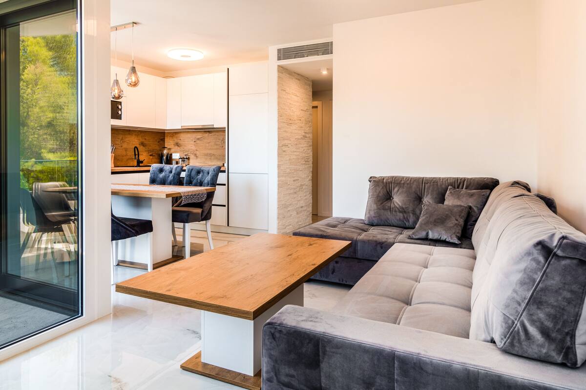 A modern living room with a grey sofa, a wooden coffee table, and a dining area with a white table and chairs.
