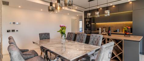 Une cuisine moderne dotée d’un îlot central, de luminaires suspendus et d’un coin repas avec une table en marbre et des chaises rembourrées.