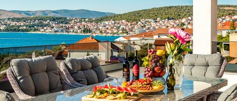 Una mesa con cubierta de vidrio, sillas de mimbre, una bandeja de frutas y una botella de vino en un balcón con vistas a un pueblo costero.