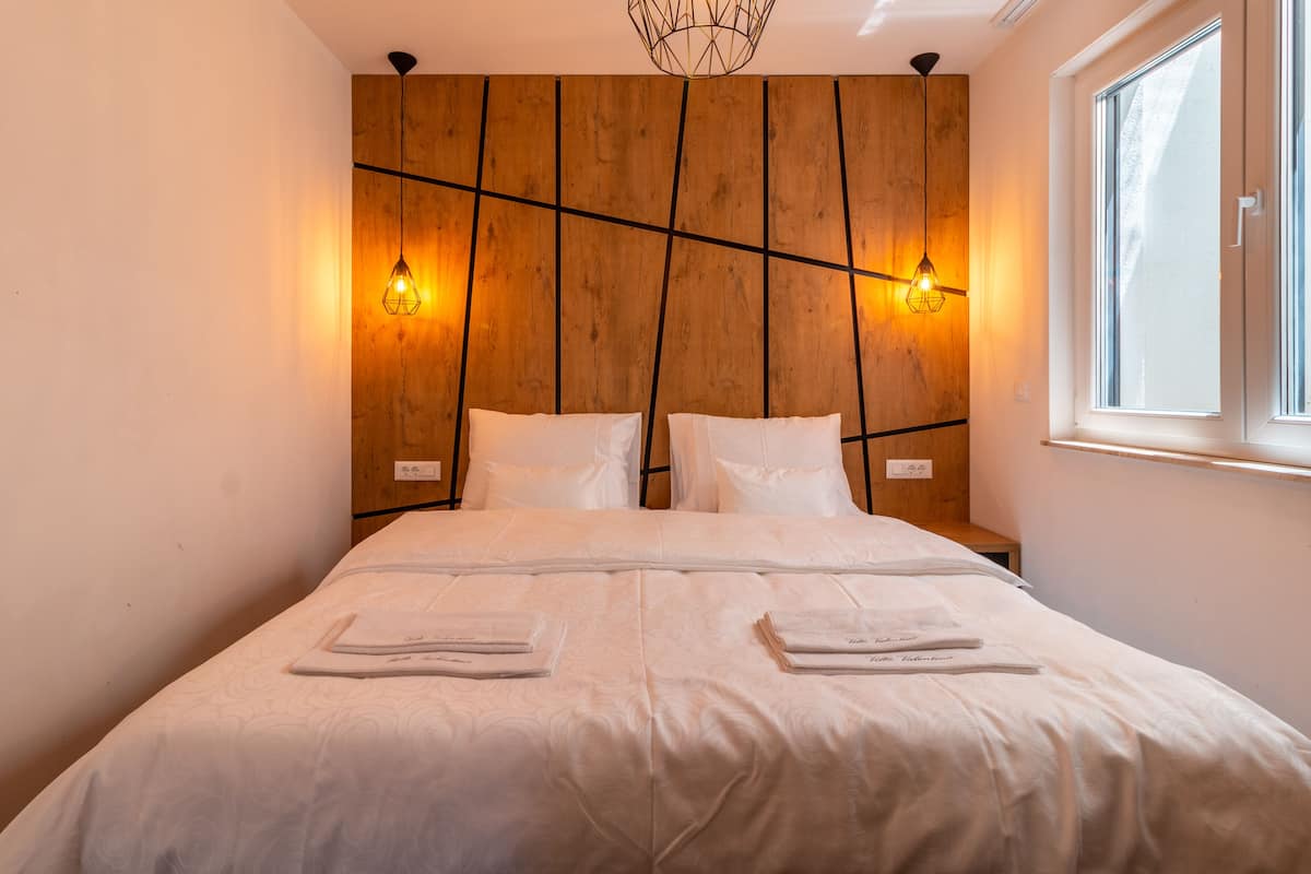 A modern hotel room with a wooden headboard, two bedside lamps, a large bed with white linens, and a window with blinds.