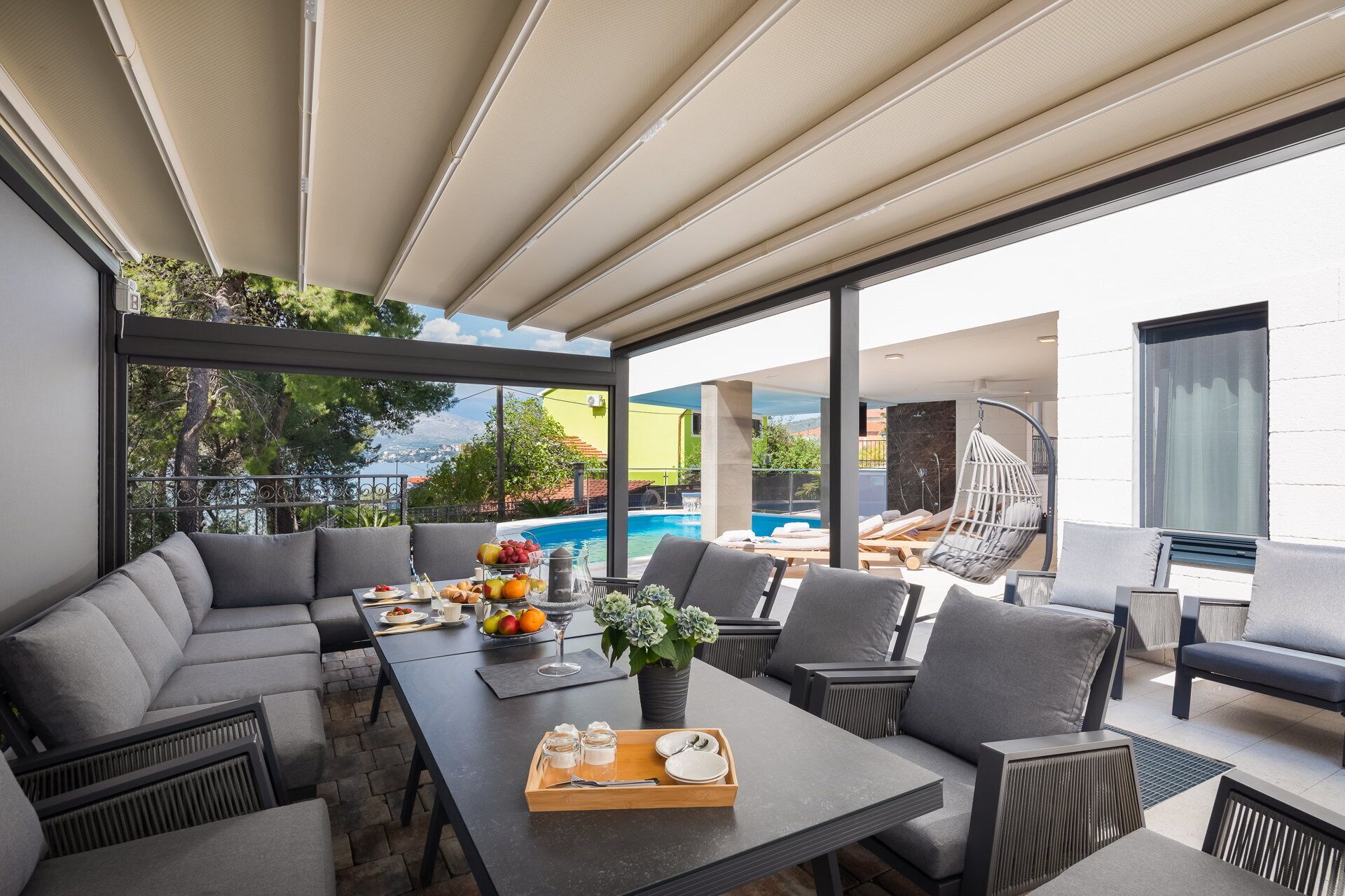 Un área de comedor moderna al aire libre con vista a la piscina, mobiliario gris y un frutero.