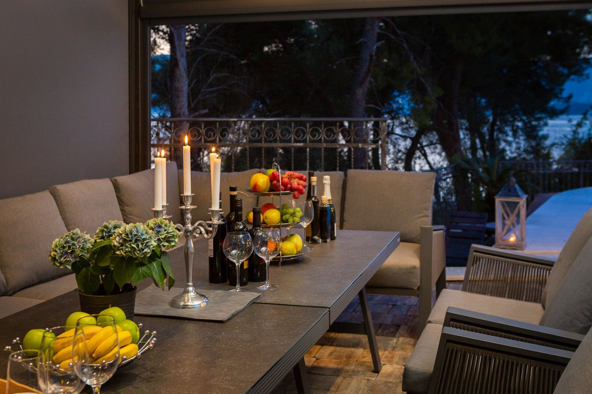 A dining table set with candles, fruits, wine glasses, and bottles, surrounded by cushioned seating.