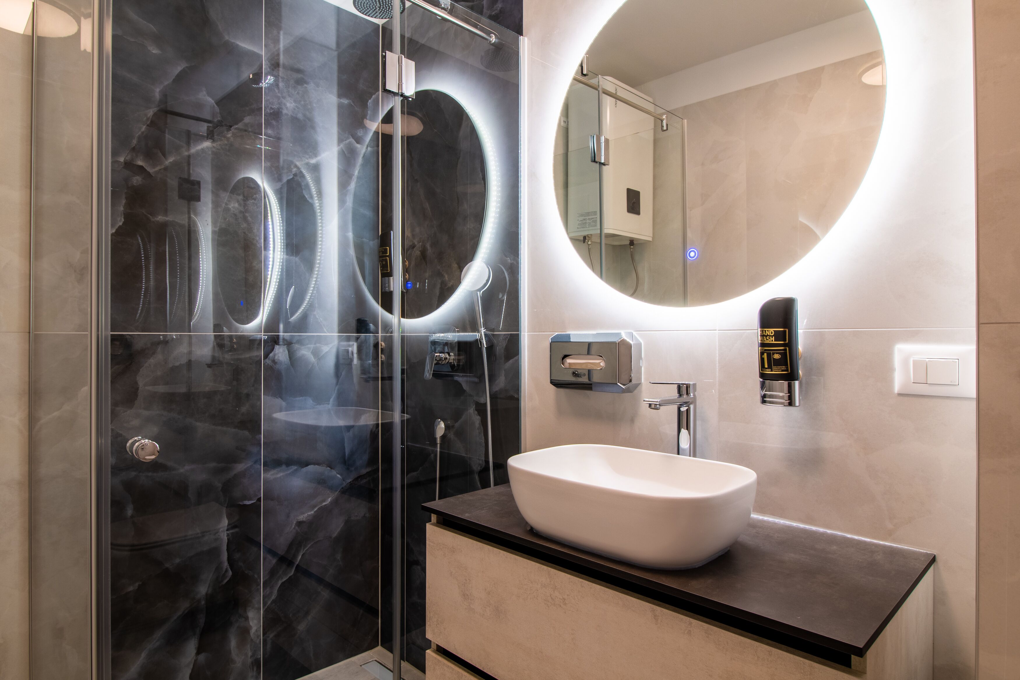 A modern bathroom with a glass-enclosed shower, a round mirror, and a white sink.
