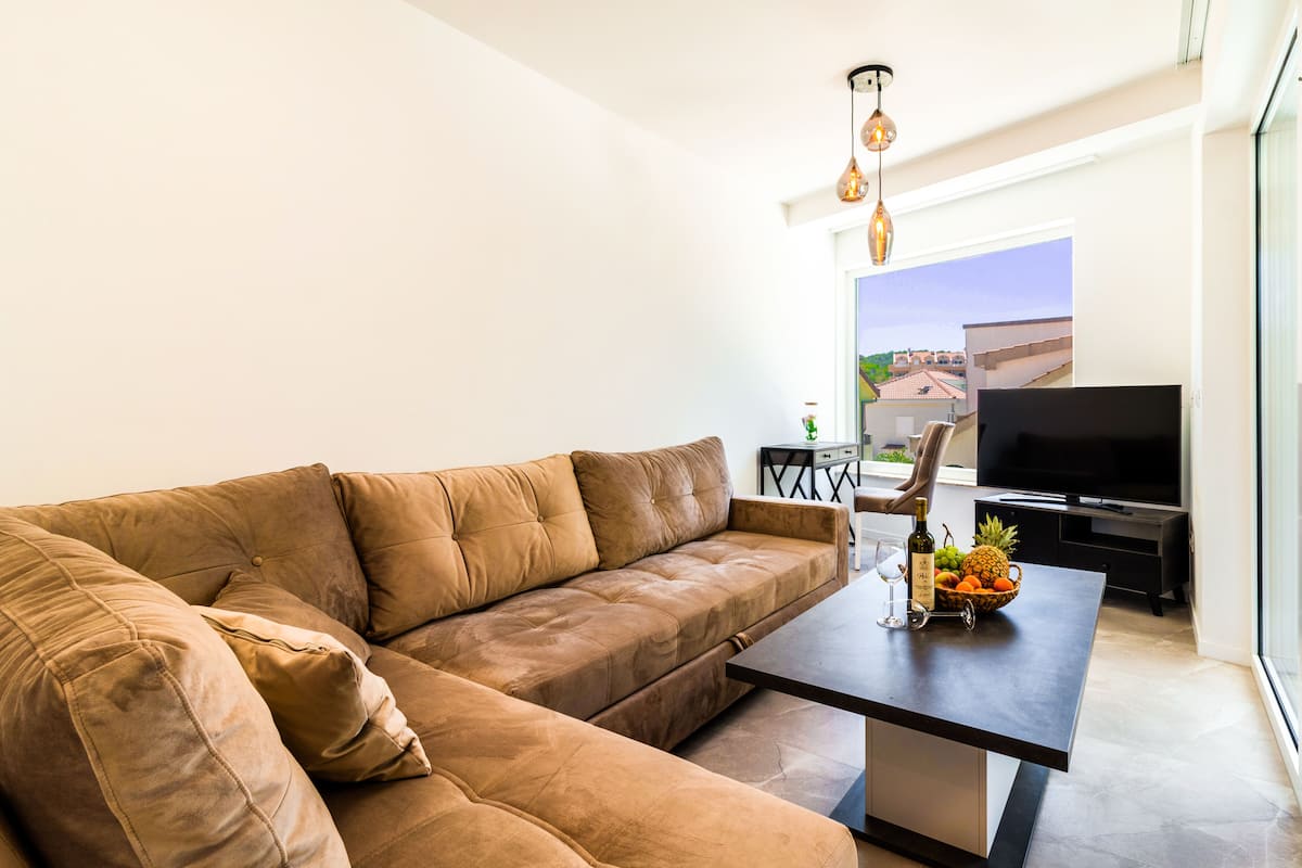 A modern living room with a leather sofa, a coffee table with fruit and a bottle, a TV, and a balcony with a view.