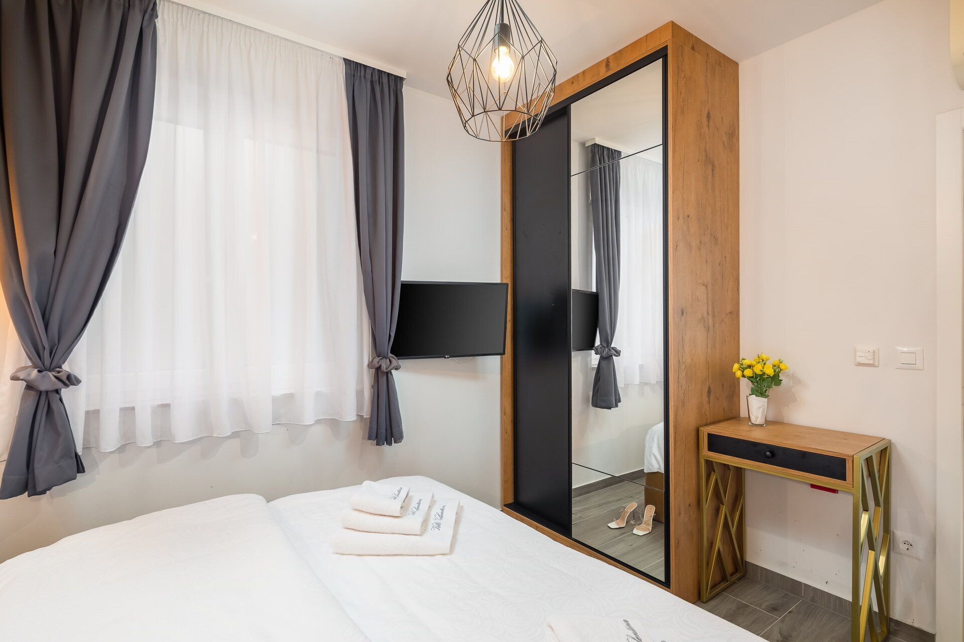 Una habitación de hotel con cama, espejo, televisor y una pequeña consola decorada con un jarrón de flores.