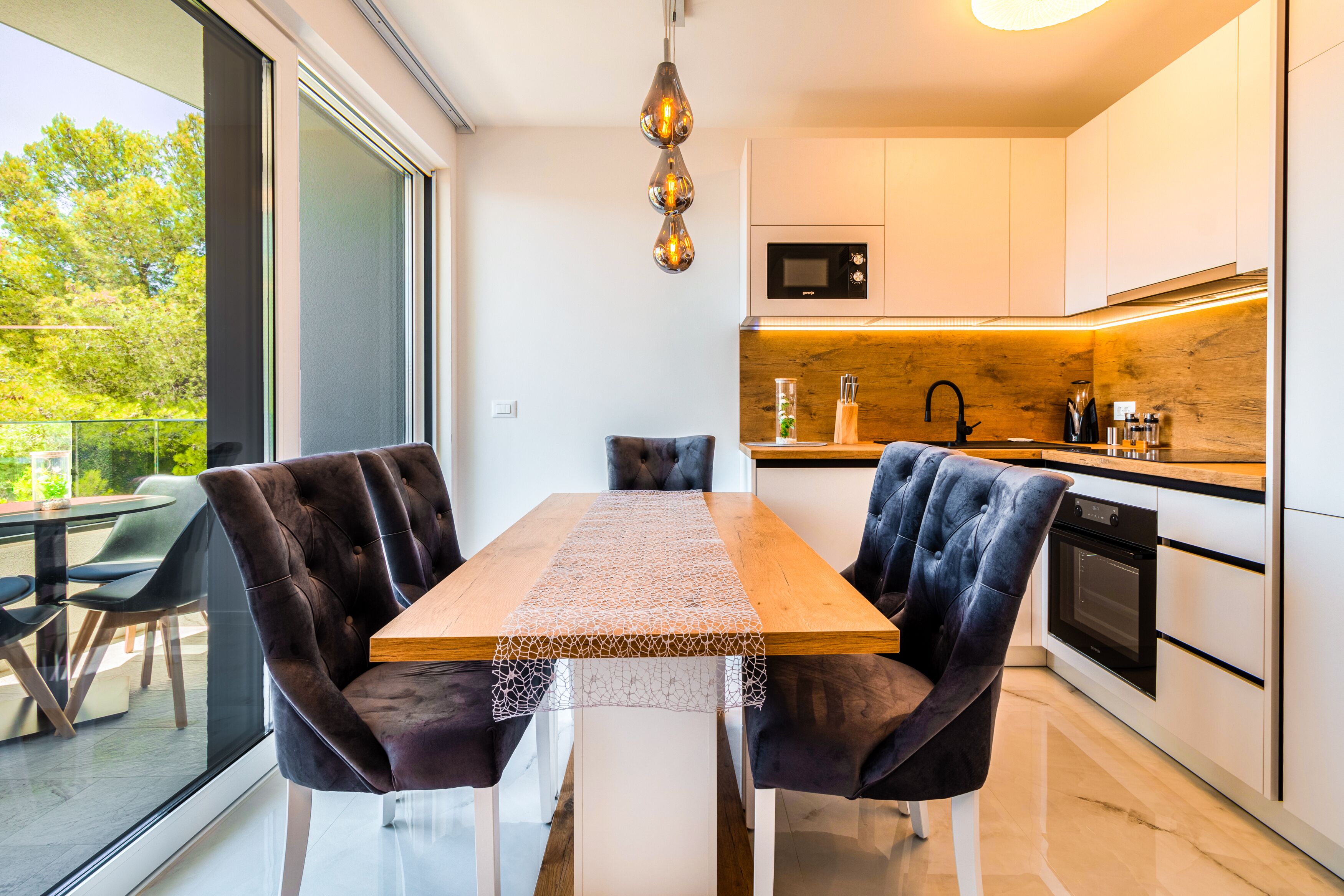 A modern kitchen with a wooden table and velvet chairs, a large window, and a view of greenery.