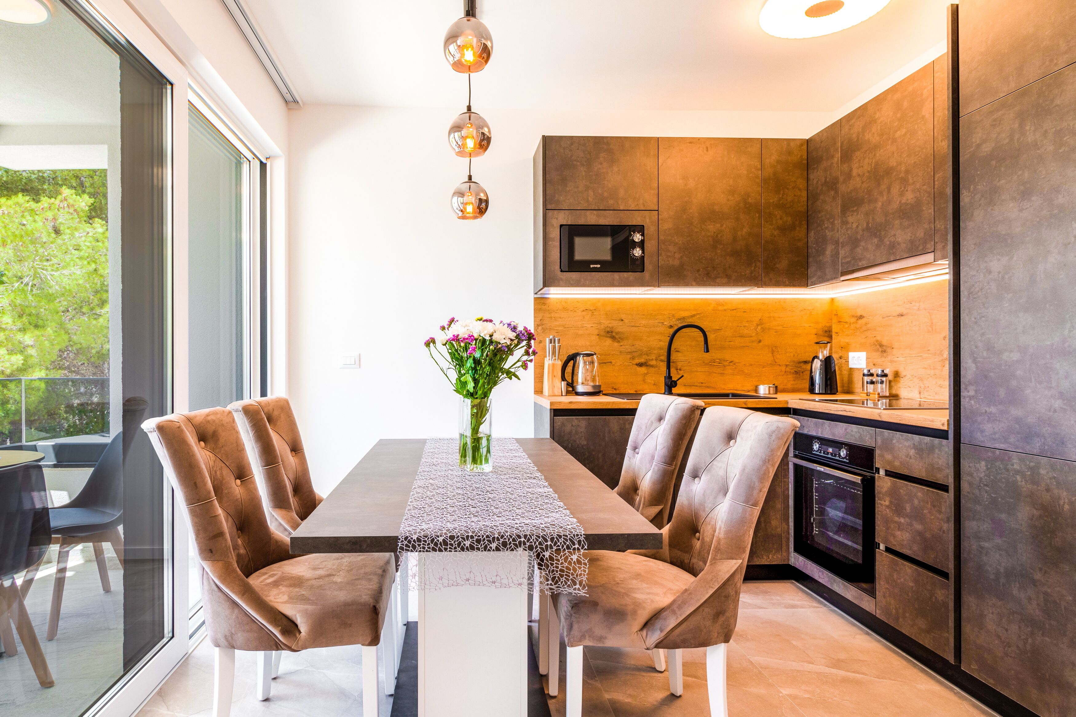 A modern kitchen with dark cabinetry, a built-in oven, and a dining area with two velvet chairs and a table.