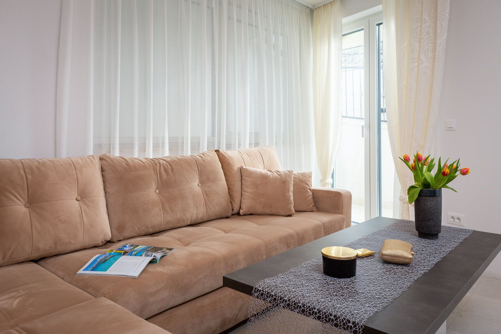 A beige sofa with a magazine, a coffee table with a vase of tulips, and a window with sheer curtains.