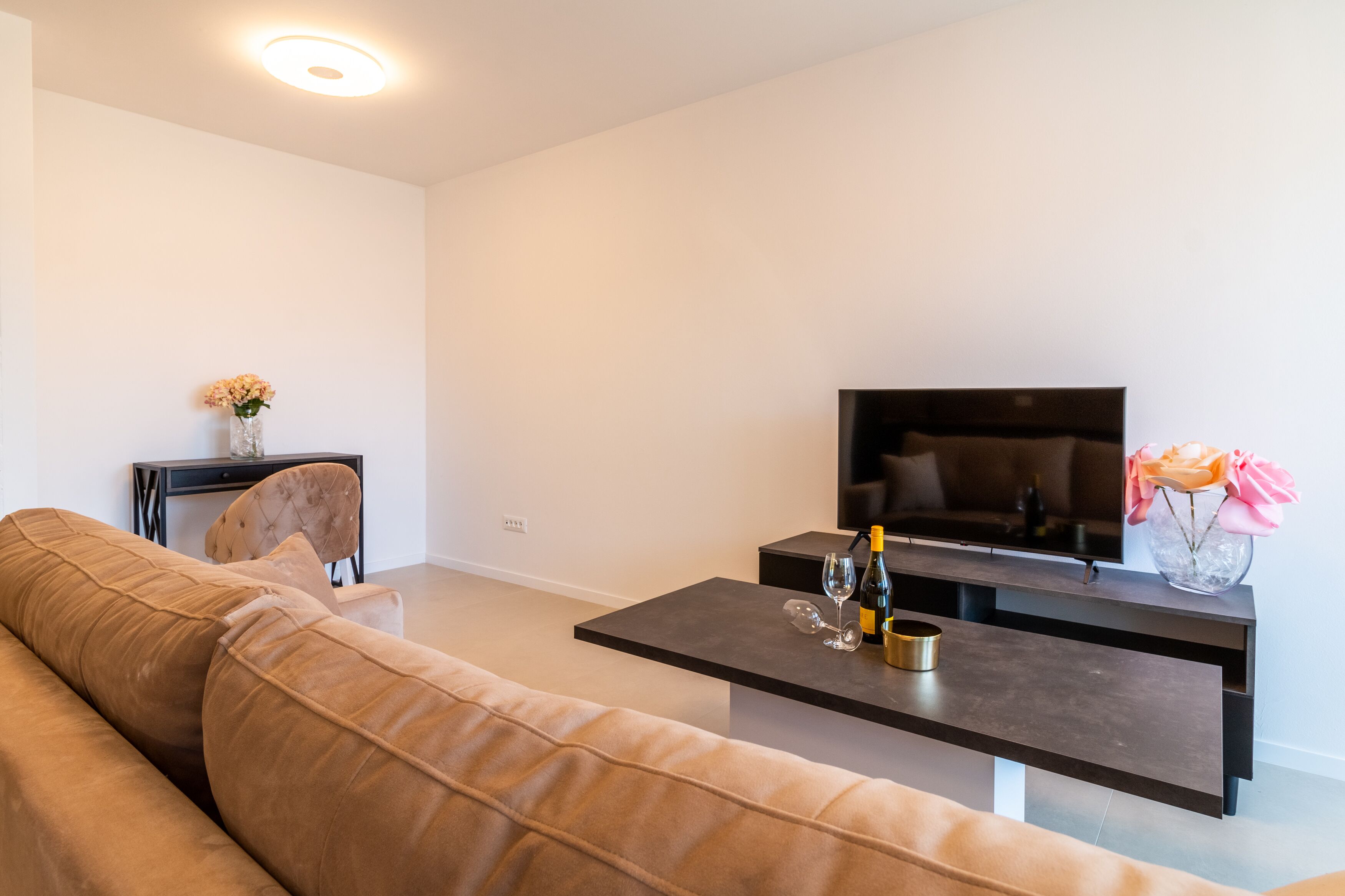 A modern living room with a sofa, a coffee table with a bottle and glasses, a TV, and a vase with flowers.