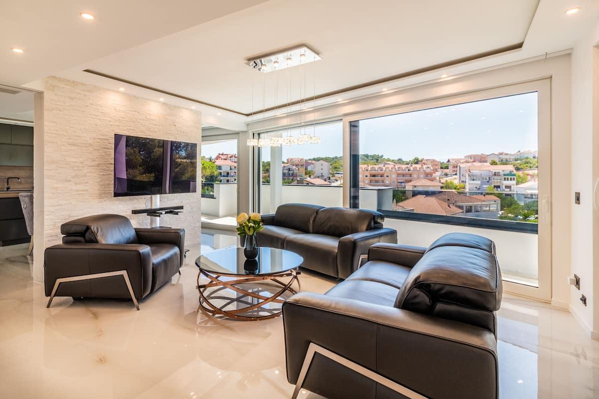 A modern living room with a large window, leather sofas, a glass coffee table, and a flat-screen TV mounted on the wall.