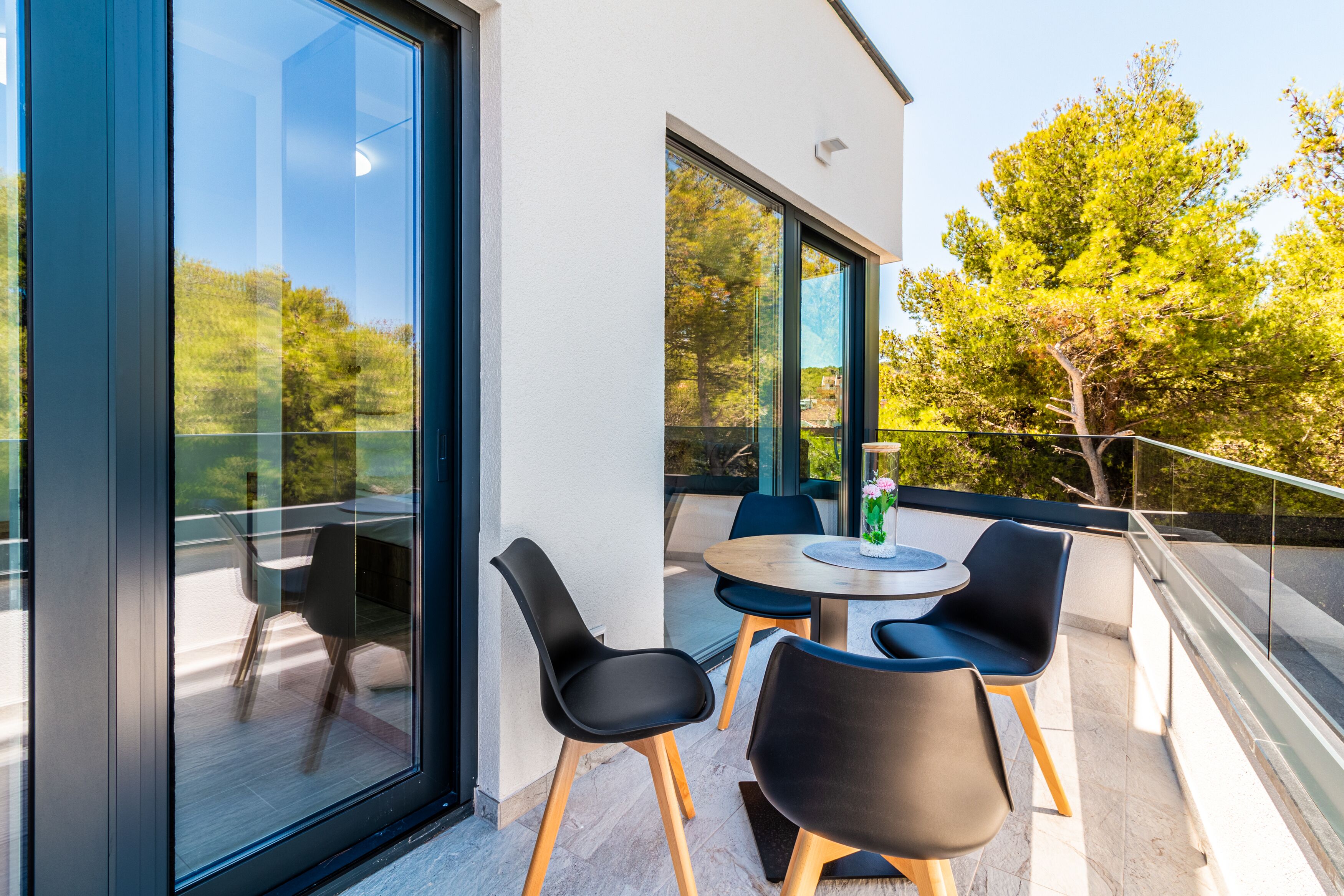 A modern outdoor seating area with a round table and four chairs, large glass windows, and a view of trees.