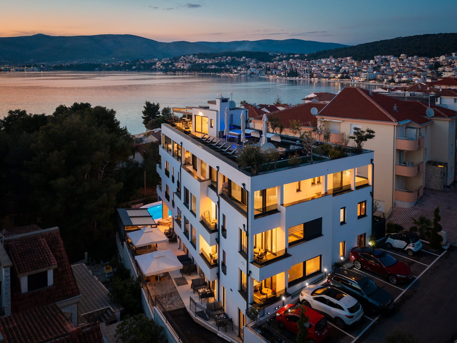 A modern multi-story hotel with balconies, outdoor seating, and a view of a lake and cityscape at dusk.
