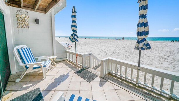 A beachside porch with two striped beach umbrellas, a white chair, and a view of the beach. - Very Merry - Renovated Gulf-Front Escape, Steps To Beach (Panama City Beach)