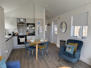 A compact kitchen with white cabinets, a stove, and a dining area with blue chairs and a wooden table. - The Lake House -  a meadow lodge that sleeps 4 guests  in 2 bedrooms (Filey)