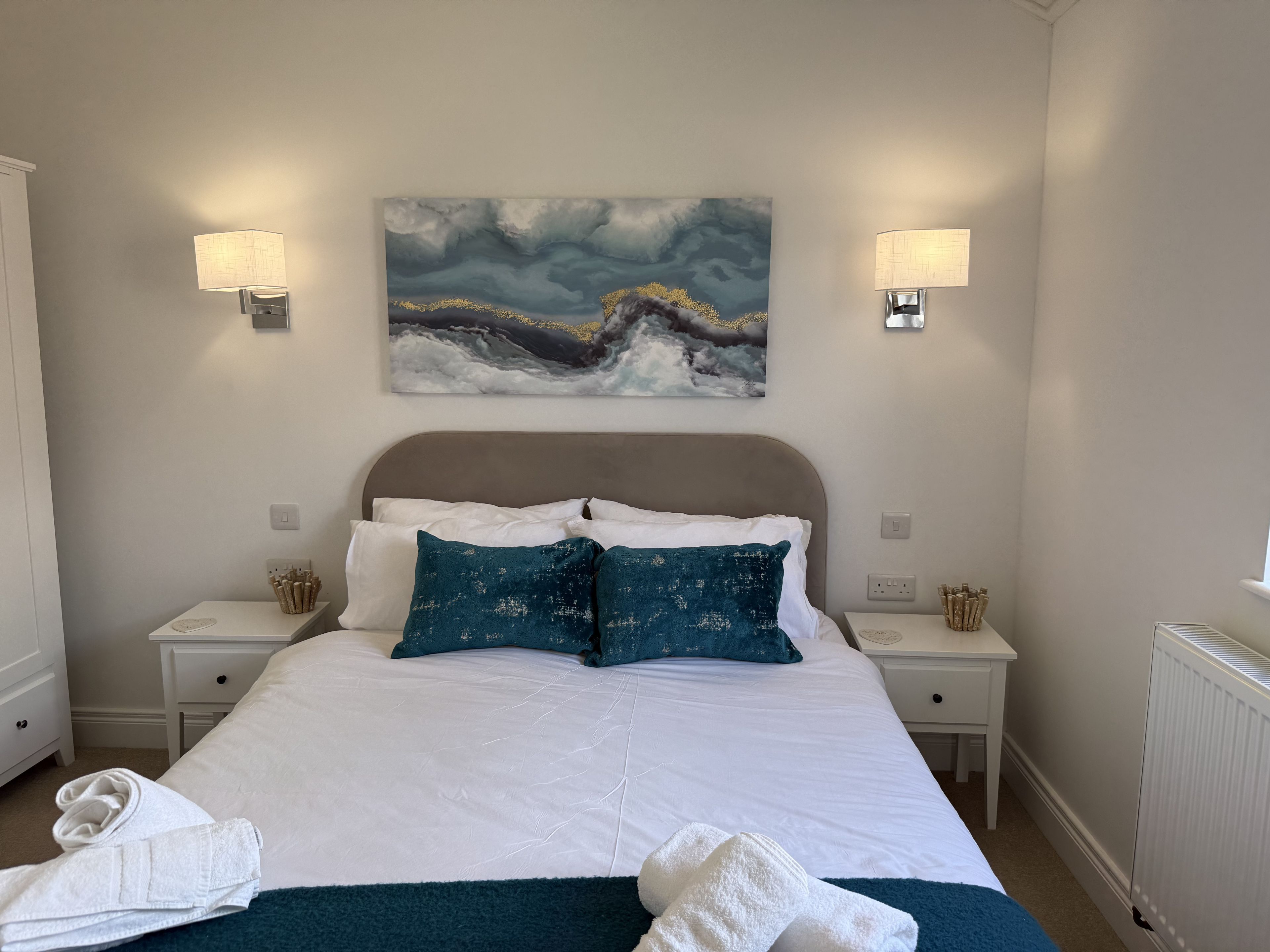 A neatly made bed with white and teal pillows, a blue throw blanket, and white towels on the floor. There are two bedside tables with lamps and a painting above the bed.