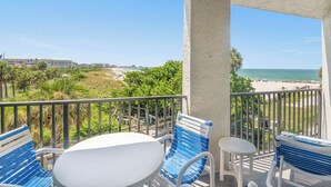 A balcony with a white table and blue chairs, overlooking a beach and greenery. - 🌴 Caprice Resort Unit #201 – Corner Gulf Front Condo with Water Views (Saint Pete Beach)