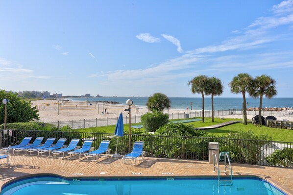 A pool area with lounge chairs, a view of the beach, and a clear sky.