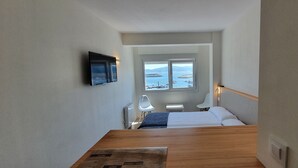 A hotel room with a bed, a desk, a chair, a TV, and a window with a view of the sea and boats. - Estudio Vista Mar Muxia (Muxía)