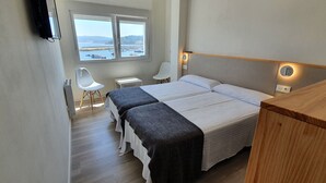 A hotel room with a large bed, a desk, a chair, and a view of the sea through two windows. - Estudio Vista Mar Muxia (Muxía)