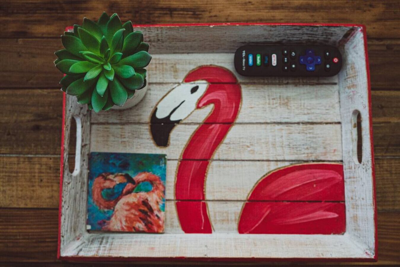 A wooden tray with a red flamingo, a remote control, and a small painting.