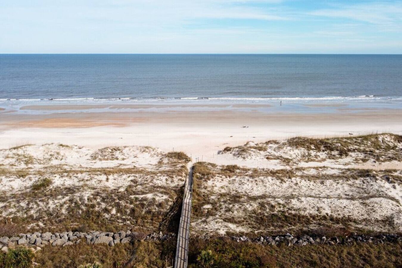 A beach with a paved walkway leading to the water, surrounded by dunes and sparse vegetation.