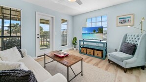 Ein Wohnzimmer mit einem Fernseher auf einem blauen Schrank, einem weißen Sofa, einem Holztisch und einem hellblauen Sessel.