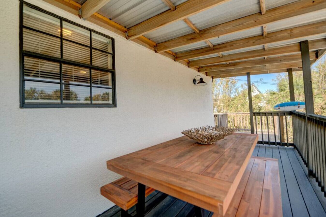 A covered outdoor area with a wooden table and benches, a railing, and a window with blinds.