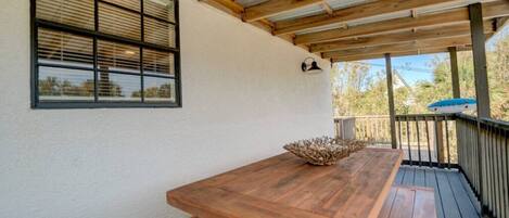 Un espacio al aire libre cubierto con una mesa de madera y bancos, barandilla y una ventana con persianas.