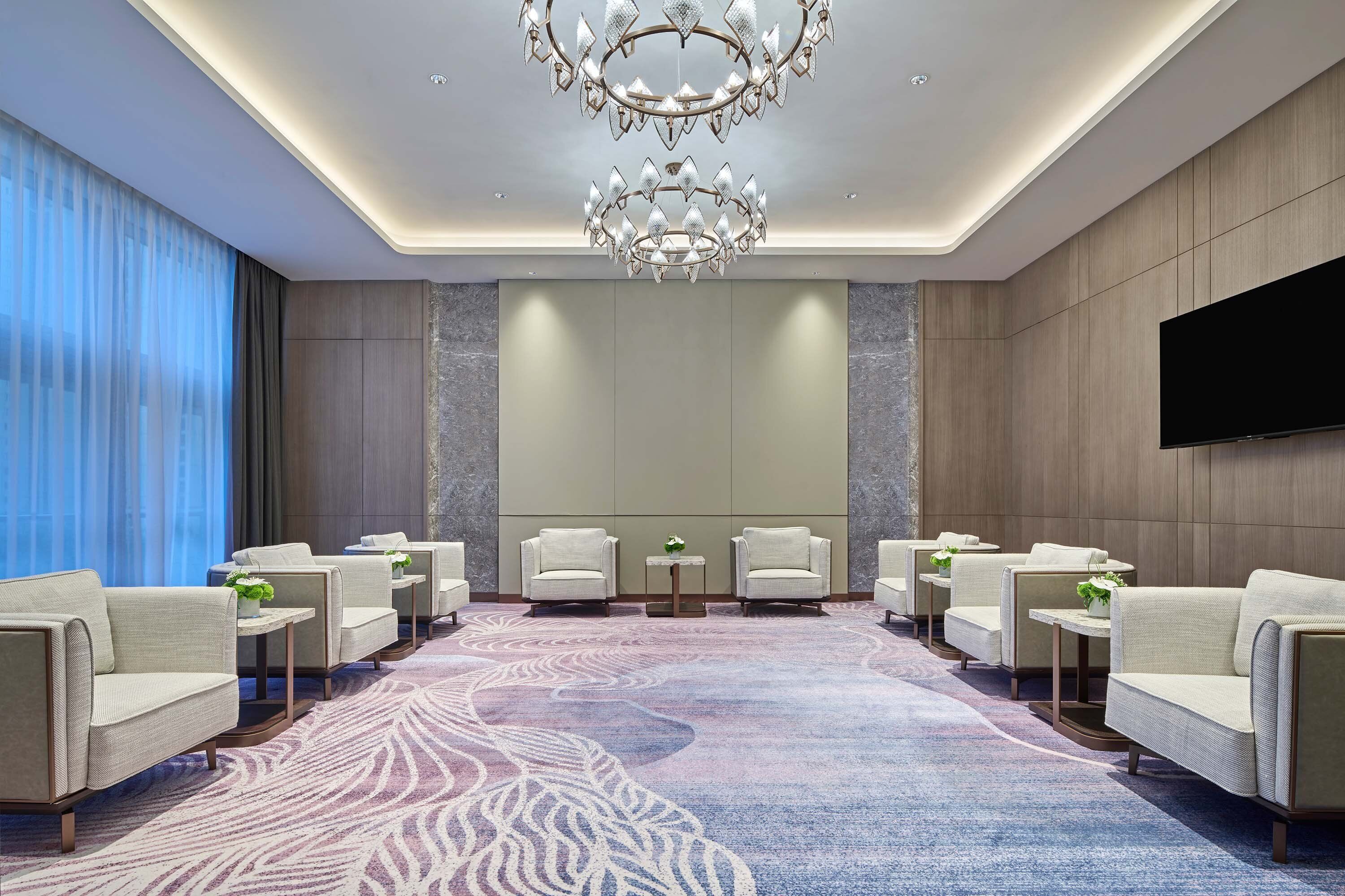 A modern hotel lobby with a large chandelier, seating area, and a flat-screen TV on the wall.