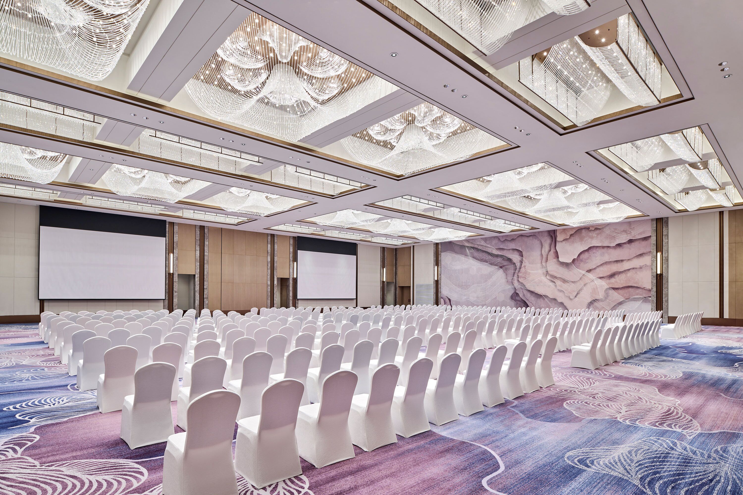 Un salón de conferencias con hileras de sillas blancas, una pantalla grande y una alfombra estampada.